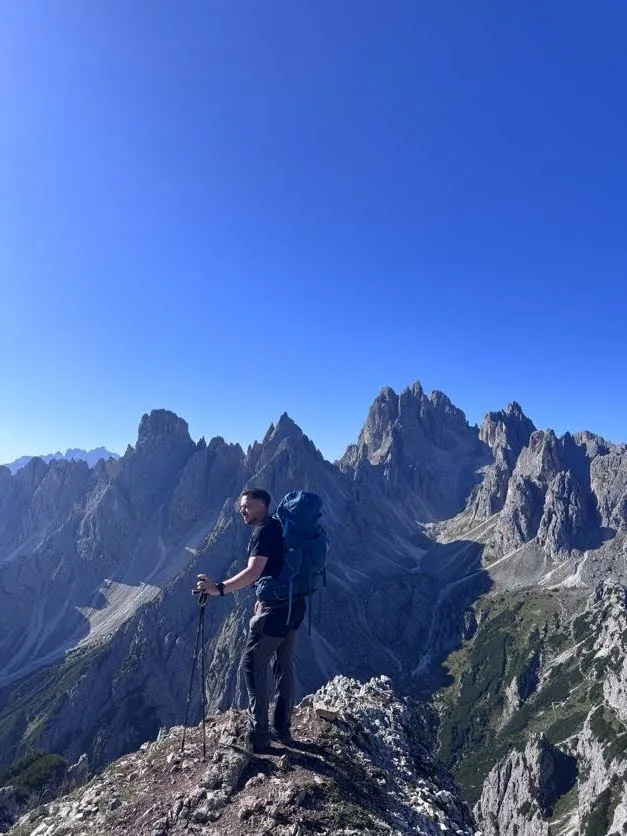 Hiking with a backpack and trekking poles on a rocky ridge beneath jagged peaks.